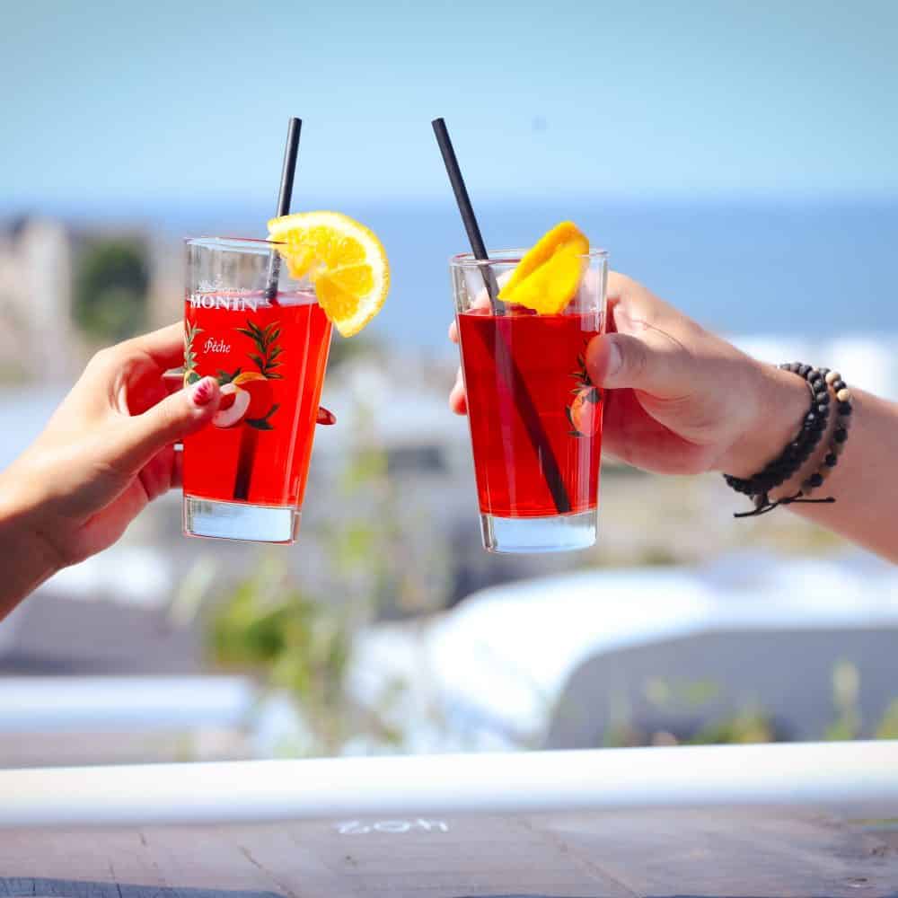 Two hands toasting red cocktails with orange slices by ocean