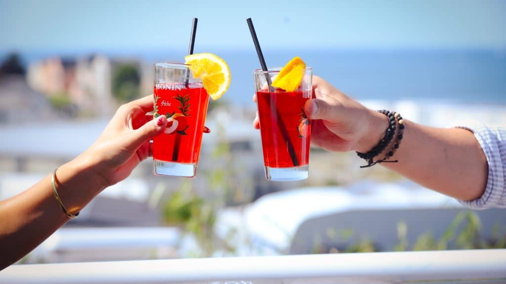 Two hands toasting red cocktails with orange slices by ocean
